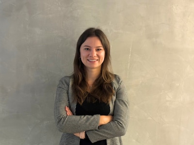 Katarina Hamann steht vor einer Wand in Betonoptik und lächelt mit verschränkten Abend in die Kamera. 