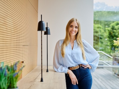 Leandra Thielemann lehnt an einer hellen Holzwand und lächelt in die Kamera.