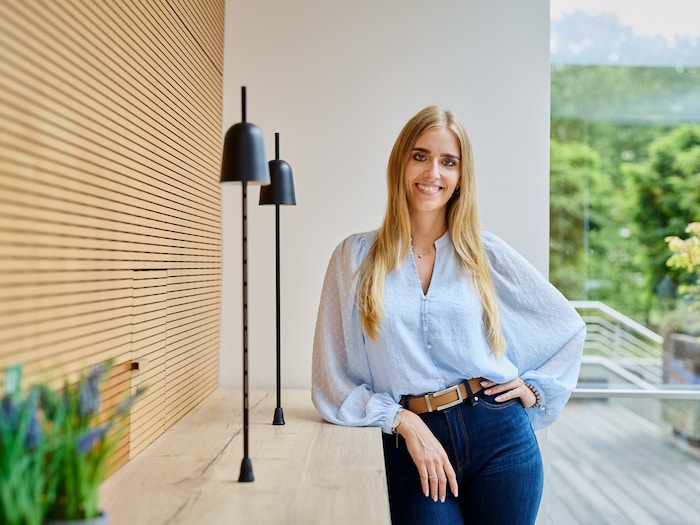 Leandra Thielemann lehnt an einer hellen Holzwand und lächelt in die Kamera.