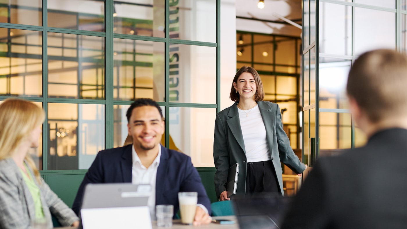 Gruppe von Personen in einem modernen Büro, im Gespräch am Tisch mit Laptops und Getränken; eine Person steht mit Laptop nahe