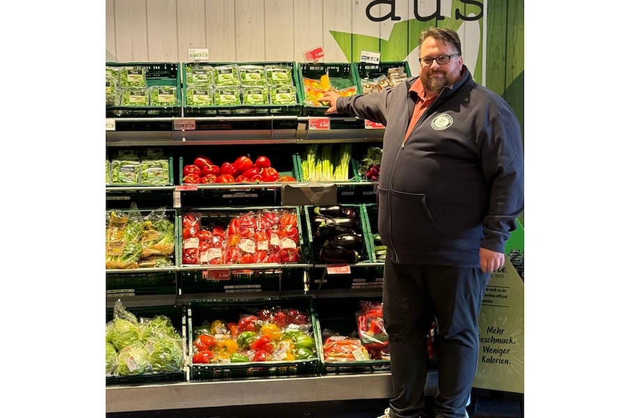 Ein Mann mit Brille steht vor dem Gemüseregal und lächelt in die Kamera.