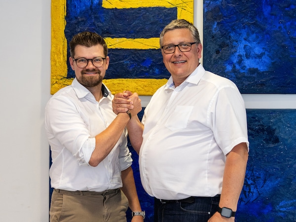 Die beiden IT-Leiter reichen sich die partnerschaftlich die Hände, im Hintergrund das EDEKA-Logo als Gemälde.