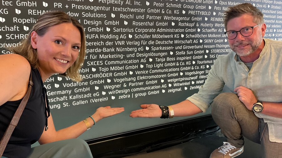 Eine Frau und ein Mann knien und zeigen auf den Namen ihrer Agentur auf einer Schautafel.