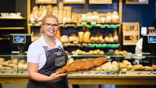 Eine Bäckereifachverkäuferin präsentiert frisch gebackene Brötchen auf einem Backblech