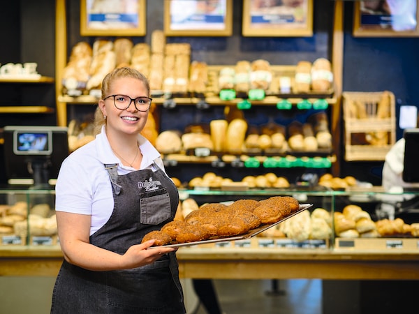 Eine Bäckereifachverkäuferin präsentiert frisch gebackene Brötchen auf einem Backblech