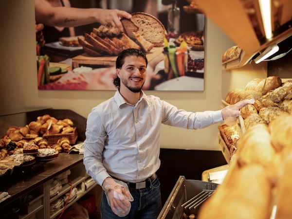 Ein junger Mann mit Plastikhandschuhen steht in der Backwarenabteilung hinter der Theke und greift nach einem Brot im Regal.