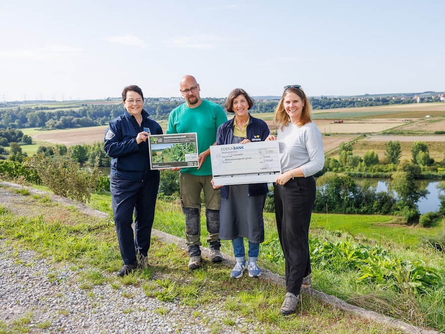 3 Frauen und 1 Mann stehen mit symbolischem Scheck vor einer grünen Landschaft mit Fluss und Feldern.
