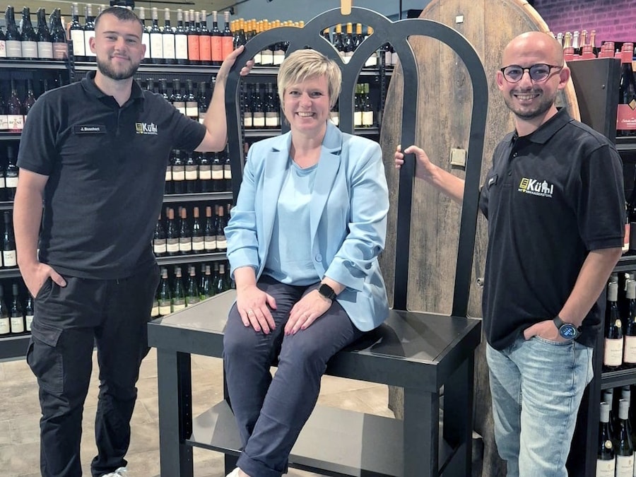 Eine Frau sitzt in der Weinabteilung des Marktes auf einem übergroßen Stuhl flankiert von 2 Männern.