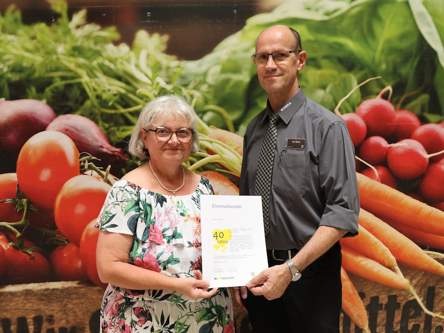 Die Jubilarin und der Marktleiter stehen mit der Urkunde vor einem überdimensionalen Bild mit Obst und Gemüse. 