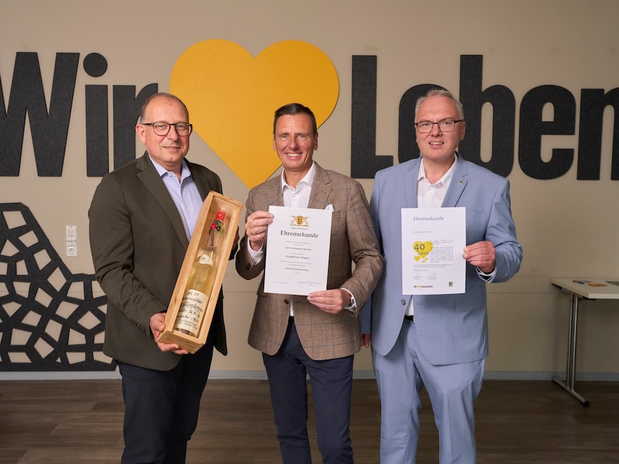 3 Männer stehen mit einem Weinpräsent und Urkunden vor einer Wand mit dem Schriftzug 