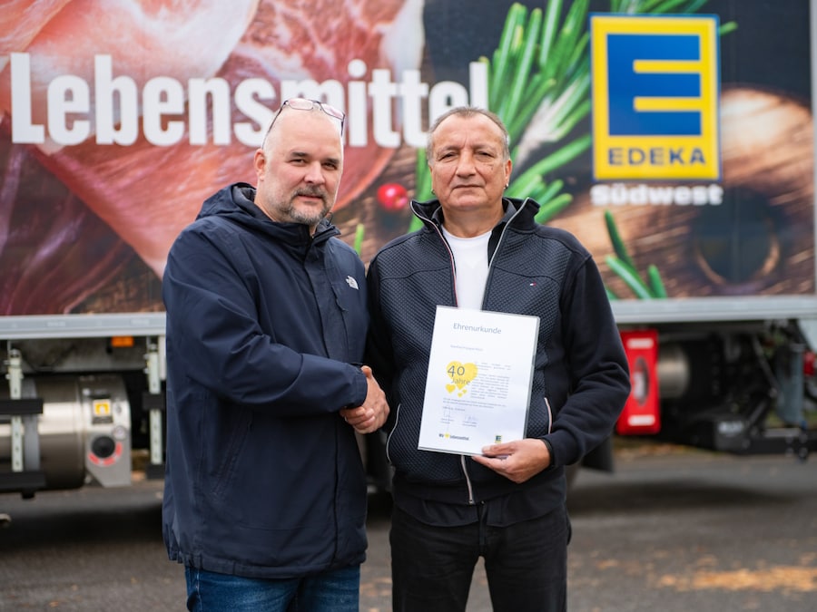 Zwei Männer stehen vor einem Lkw. Der linke gratuliert per Handschlag dem Jubilar, der eine Urkunde für 40 Jahre Treue hält.
