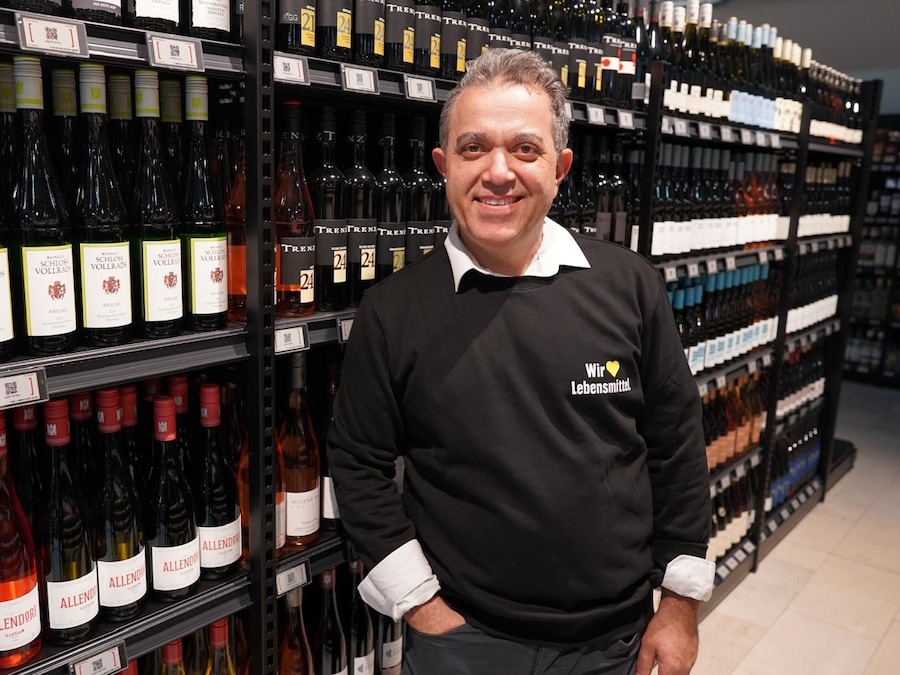 Marktinhaber Jafar Graf steht in Getränkeabteilung vor dem Weinregal und lächelt freudig.