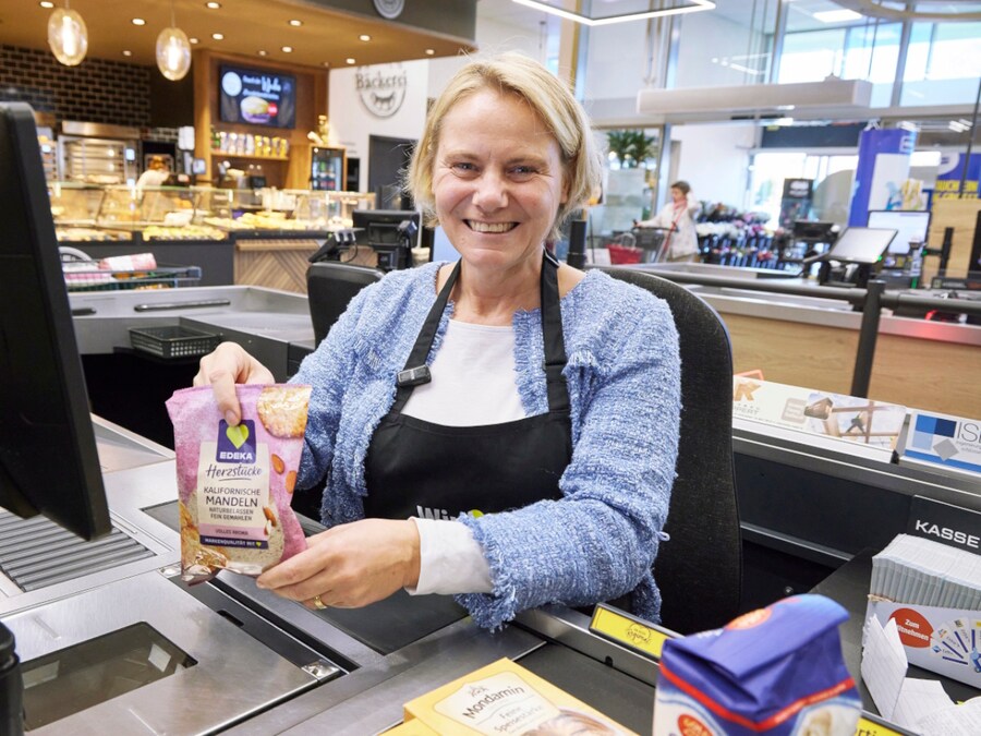 Eine Frau sitzt an der Kasse im EDEKA-Markt und lächelt. Dabei zieht sie Waren eines Kundeneinkaufs über den Scanner.