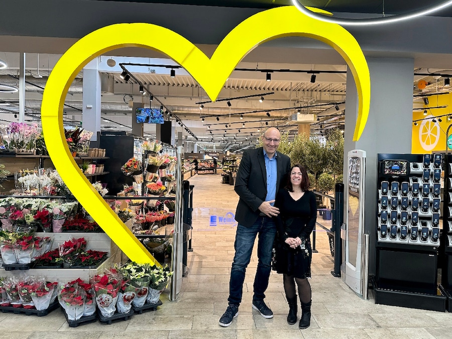 Ein Mann und eine Frau stehen vor im Eingang des Marktes vor einem großen gelben Herz.