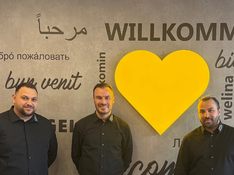3 Männer stehen vor einem Wand mit gelben Herz und Willkommensgrüßen.