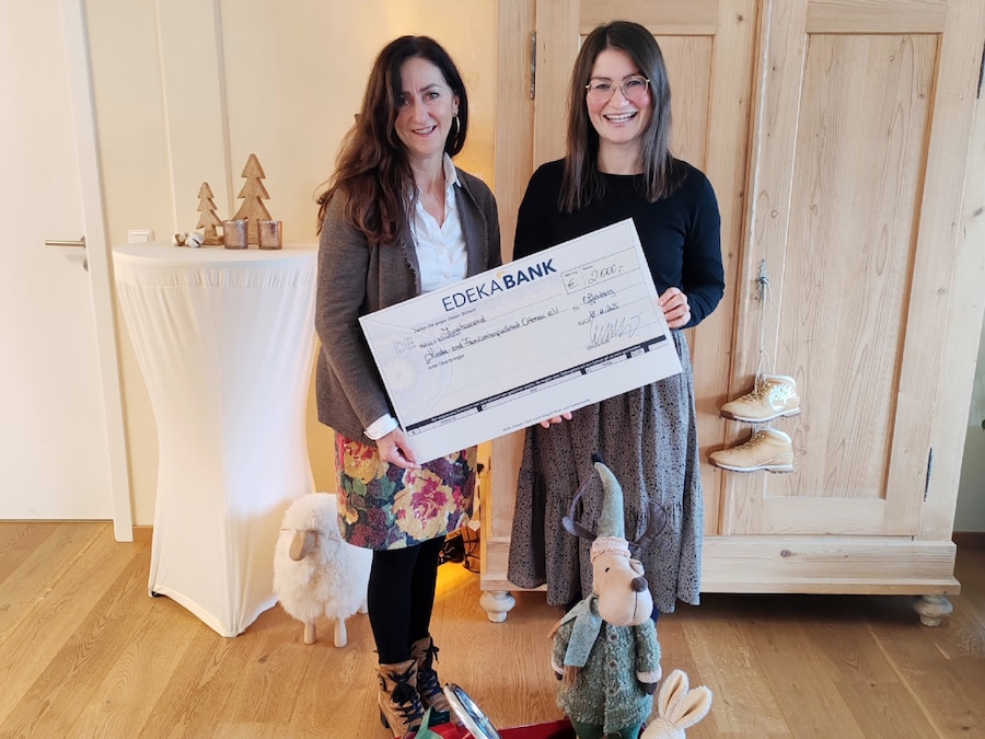2 Frauen stehen mit einem symbolischem Spendenscheck in einem Kinderspielzimmer.