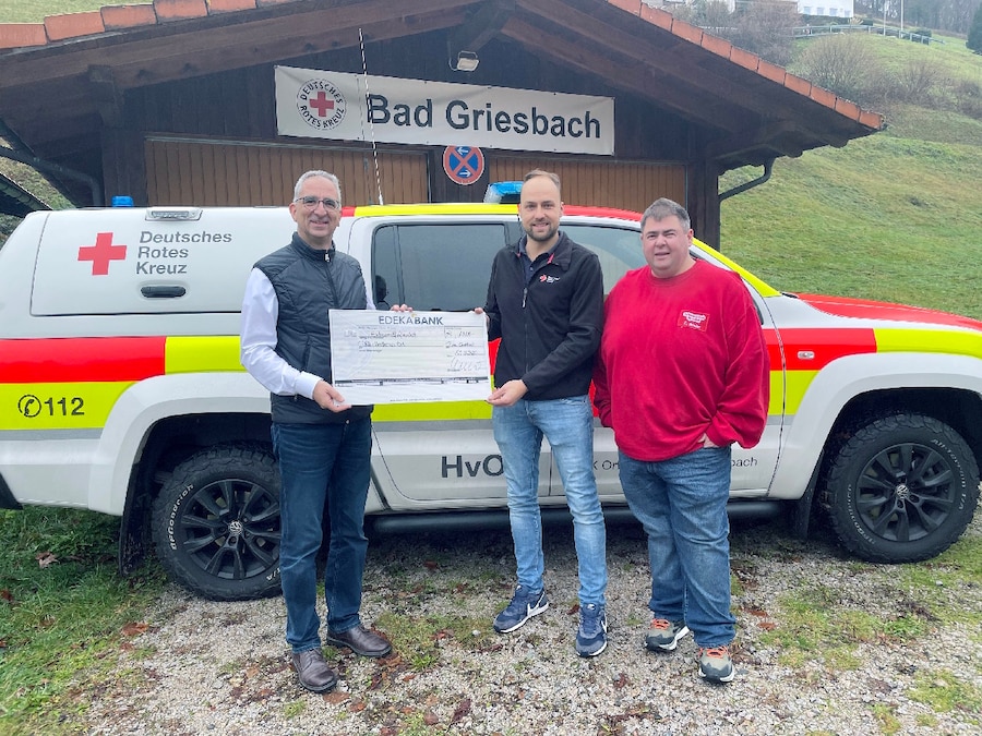 3 Männer stehen mit einem symbolischen Spendenscheck vor einer Hütte und einem DRK Auto.