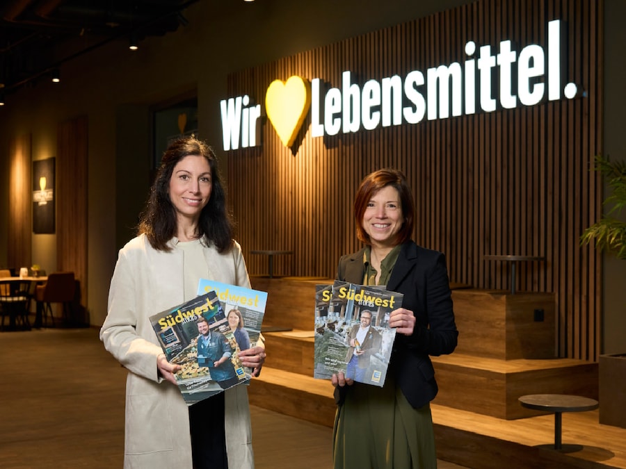 2 Frauen stehen mit dem Mitarbeitermagazin vor einer Wand mit EDEKA Logo.