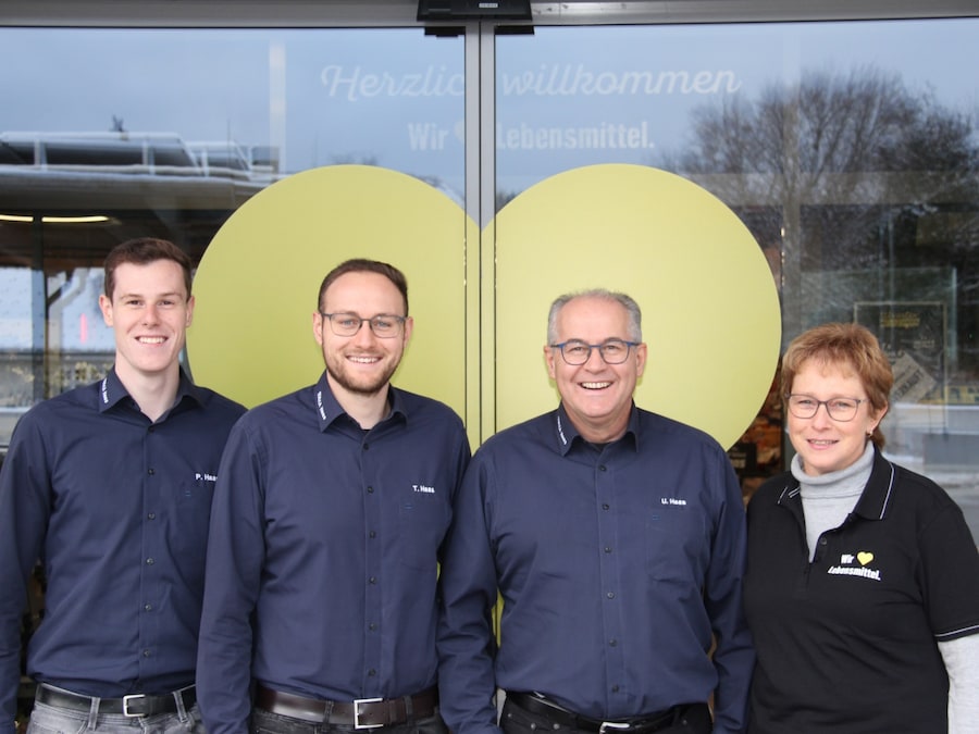 3 Männer und eine Frau stehen vor dem Eingang ihres Marktes. Auf den Schiebetüren ist ein großes gelbes Herz.