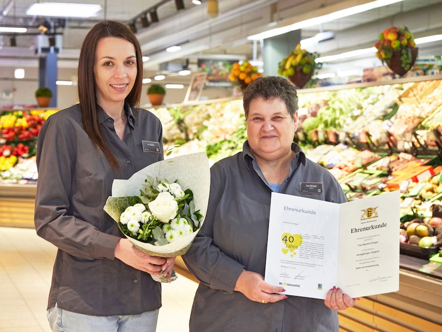 2 Frauen stehen im Markt vor der Obstabteilung mit einer Urkunde und einen Blumenstrauß in der Hand.