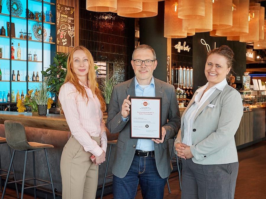Ein Mann mit Urkunde steht in der Mitte zweier Frauen vor dem Betriebsrestaurant.