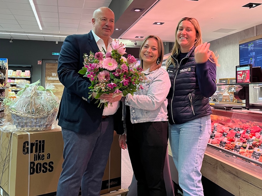 Ein Mann steht mit 2 Frauen und einem Blumenstrauß in der Hand vor der Frischetheke im Markt.