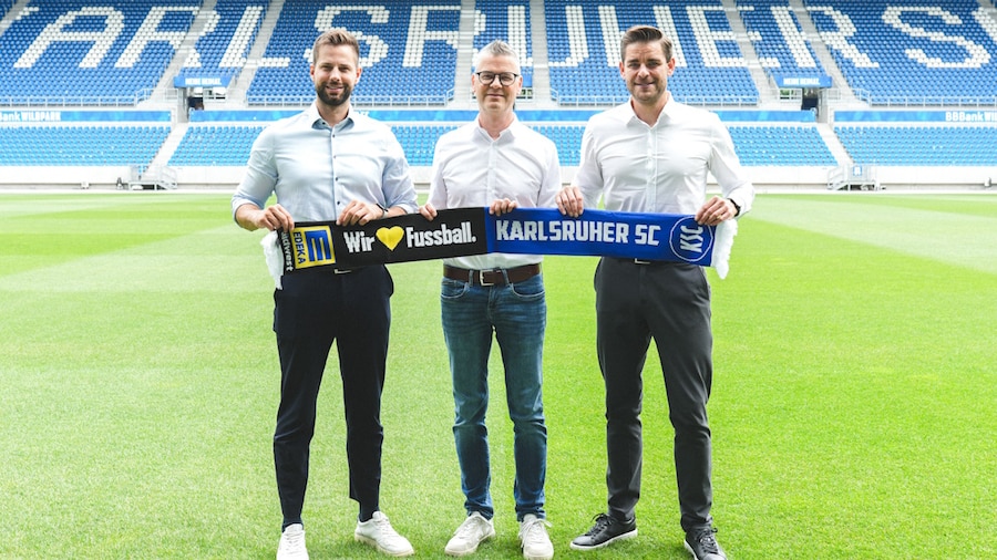 3 Männer stehen auf dem Rasen eines Fussballstadions und halten einen 