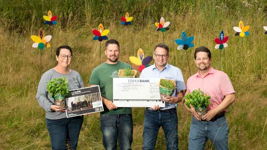Eine Frau und 3 Männer stehen mit einem symbolischem Scheck und Kräutertöpfen auf einer Wiese.