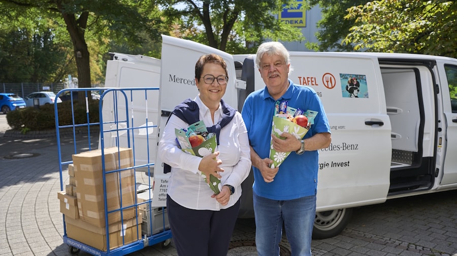 Eine Frau und ein Mann stehen vor einem Transporter und einen Rollcontainer. In der Hand halten sie gefüllte Schultüten.