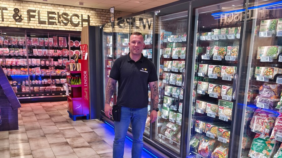Ein EDEKA-Kaufmann steht vor einem Kühlregal mit Glastüren. 