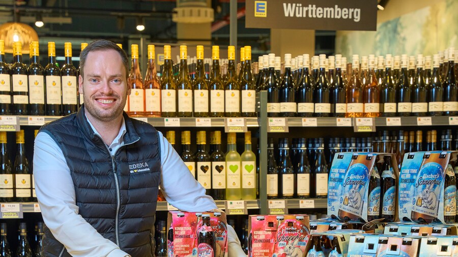 Ein EDEKA-Kaufmann lächelt vor einem Regal mit Weinen aus Württemberg. Im Vordergrund werden Bier-Sixpacks angeboten.