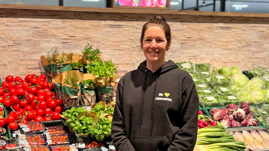 Eine junge EDEKA-Kauffrau steht lächelnd vor bunter Gemüseauslage mit frischen Produkten.