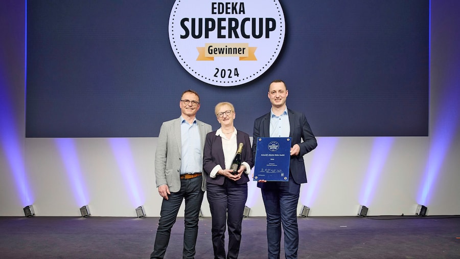 Eine Frau und zwei Männer zeigen ihre Sieger-Urkunde auf einer Bühne vor einer Leinwand mit dem Text EDEKA Supercup Gewinner