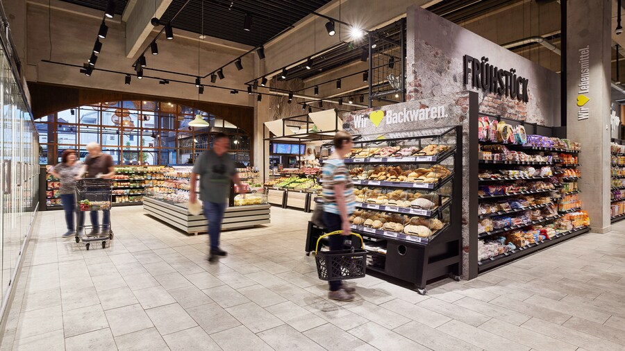 Frische-Bereich mit Obst & Gemüse und Backwaren sowie vorbeigehende Kunden mit Einkaufswägen und -körben.