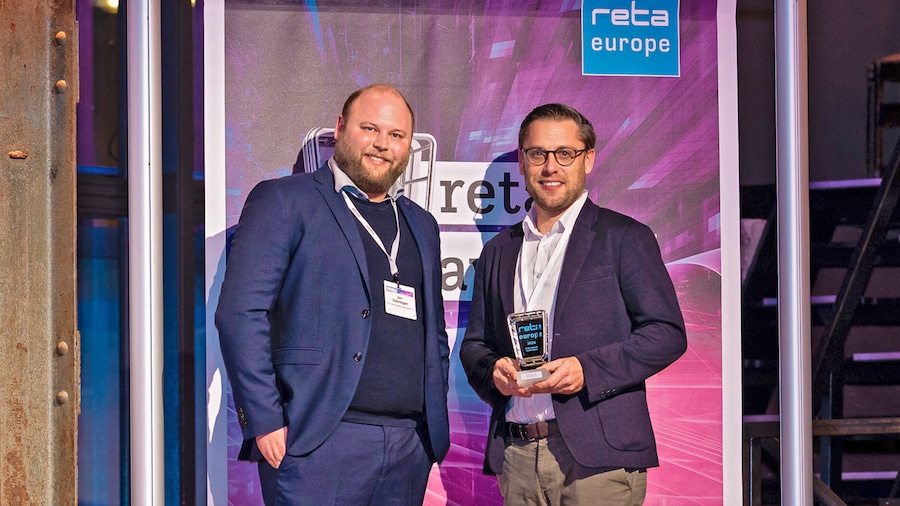 Zwei Männer posieren mit Preistrophäe vor dem Banner der reta europe.