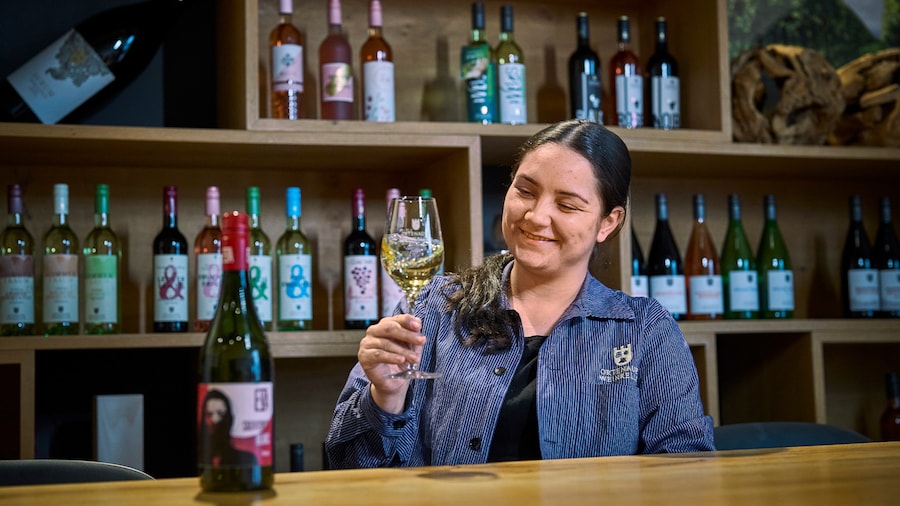Eine lächelnde Frau schwenkt ein Glas Weißwein. Die passende Weinflasche steht geöffnet auf dem Tisch.