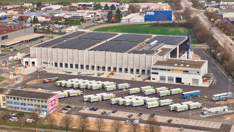 Luftaufnahme der Tiefkühllager-Baustelle mit einer großen Photovoltaikanlage auf dem Dach und vielen Lkws auf dem Parkplatz.