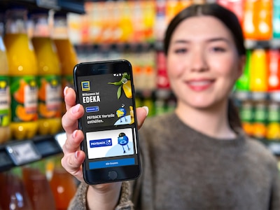 Lächelnde Frau im Supermarkt hält Smartphone mit PAYBACK-App in die Kamera.