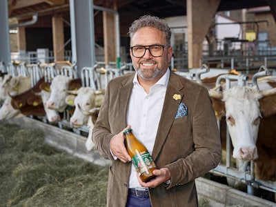 Lächelnder Mann im Sakko steht in einem Kuhstall und hält eine Flasche mit Bio-Milch. 
