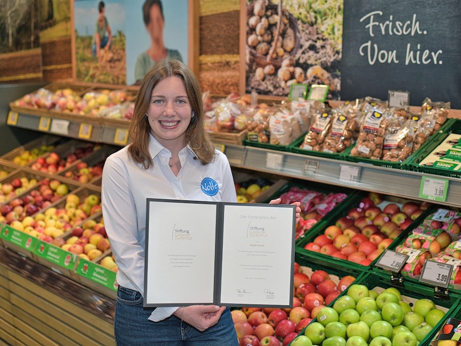 Junge Markt-Mitarbeiterin präsentiert lächelnd eine Urkunde in der Obst- und Gemüseabteilung. 
