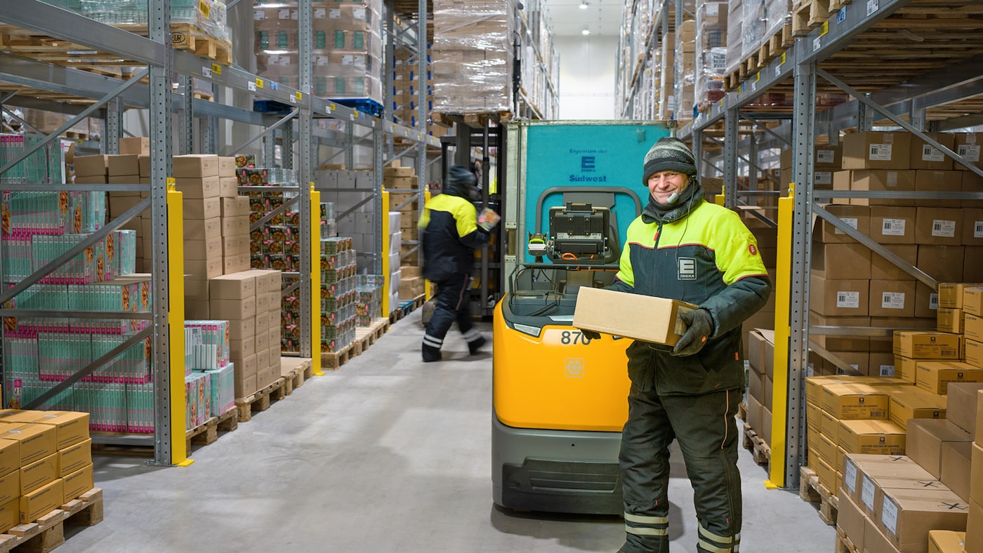 Lächelnder Mann in Arbeitskleidung mit Paket in einem Tiefkühllager, im Hintergrund ein Mitarbeiter mit Gabelstapler.