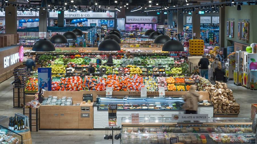 Blick durch das Innere eines Supermarkts mit Obst- und Gemüseabteilung und andere Regalbereiche im Hintergrund. 