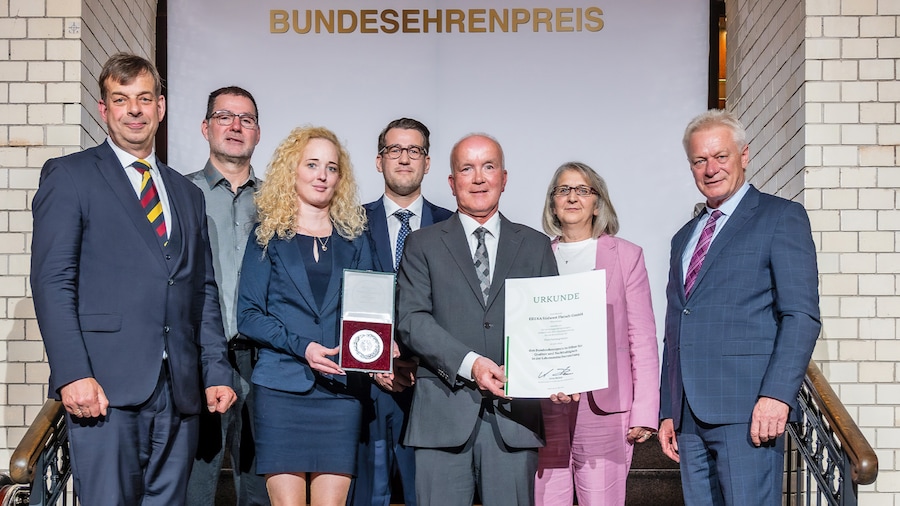 Gruppenfoto von sieben Personen auf einer Treppe, die eine Urkunde und einen Preis präsentieren. 