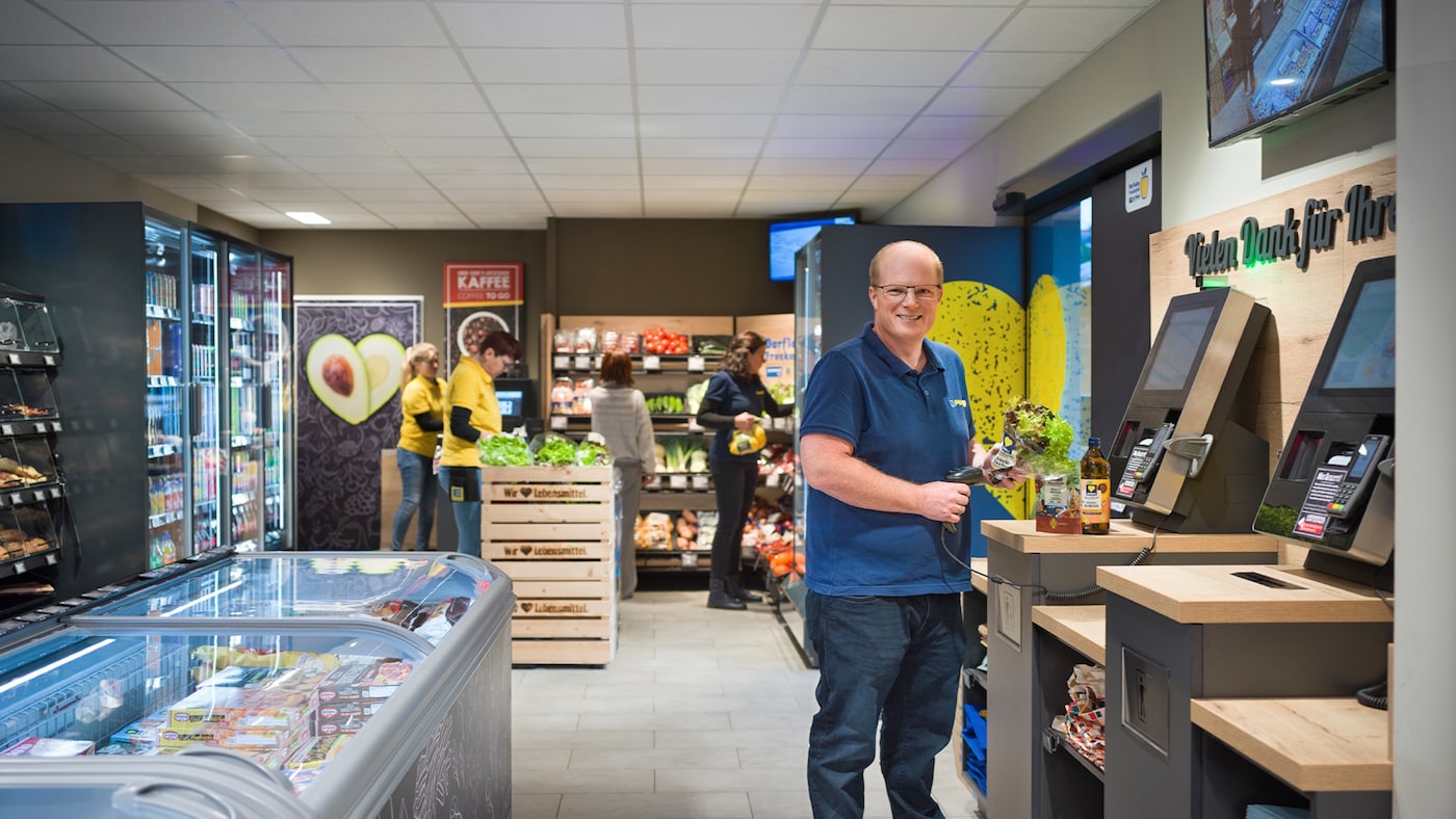 Freundlich lächelnder EDEKA-Kaufmann scannt Produkte an einer Self-Check-out-Kasse. 