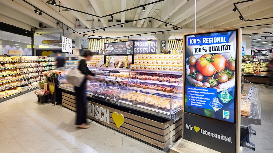 Eine Kundin begutachtet das Angebot einer Kühltheke, neben der auf einer Tafel Werbung für regionale Produkte zu sehen ist