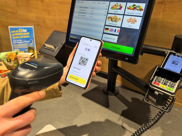 Zwei Hände scannen den QR-Code auf einem Smartphone an einer Self-Check-out-Kasse. 