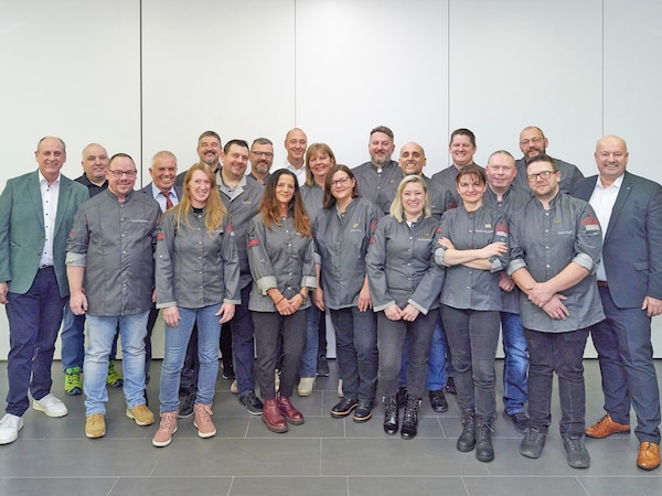 Gruppenfoto mit vielen Menschen in grauer Metzgerei-Arbeitskleidung und zwei Männer im Anzug. 