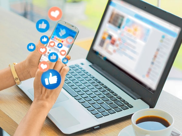 Hände halten ein Smartphone mit herausspringenden Social-Media-Reaktionen vor einem aufgeklappten Laptop 