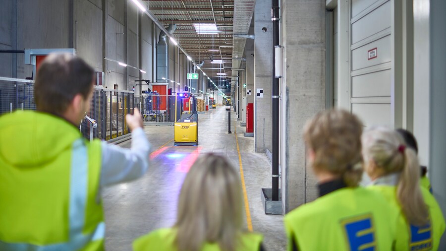 Ein Mitarbeiter zeigt einer mit Warnwesten gekleideten Besuchergruppe den Ablauf im Lager.