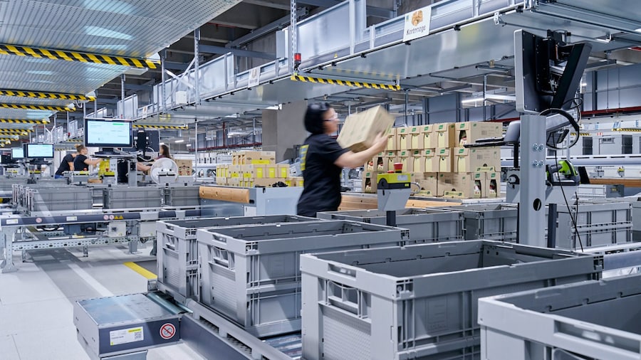 Betriebsbesichtigung: Auf einem Förderband wandern zahlreiche Klappkisten und Frauen beladen diese mit Paketen.